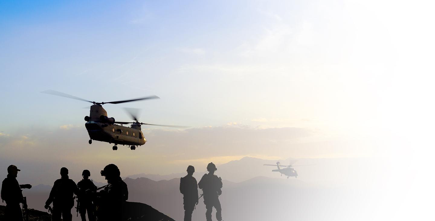 Two helicopters flying over mountains at dusk with warfighters watching