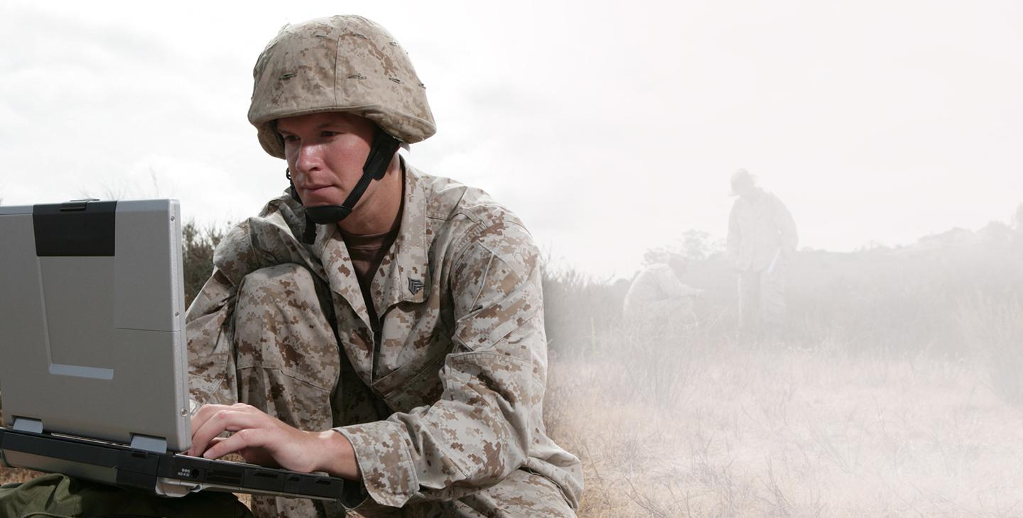 Man wearing combat gear kneeling in a field while typing on a military laptop 