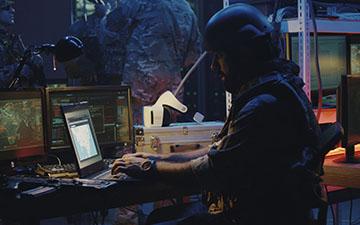 一名身穿战斗装备的男子坐在一台小型笔记本电脑前，在战场上使用Viasat服务