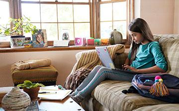 一个棕色长发的年轻女孩坐在沙发上，背包旁，用笔记本电脑做作业