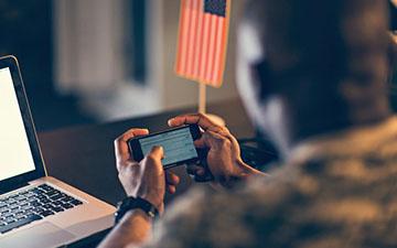 一个军人的特写镜头，他把智能手机横拿在笔记本电脑和美国国旗前面