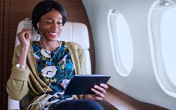 一头黑发的职业女性坐在私人飞机上, 戴着耳机在平板电脑上听内容