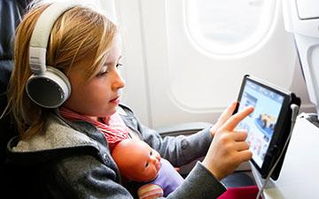 年轻的金发女孩坐在飞机上, 抱着娃娃，戴着耳机, 平板电脑上的流媒体内容
